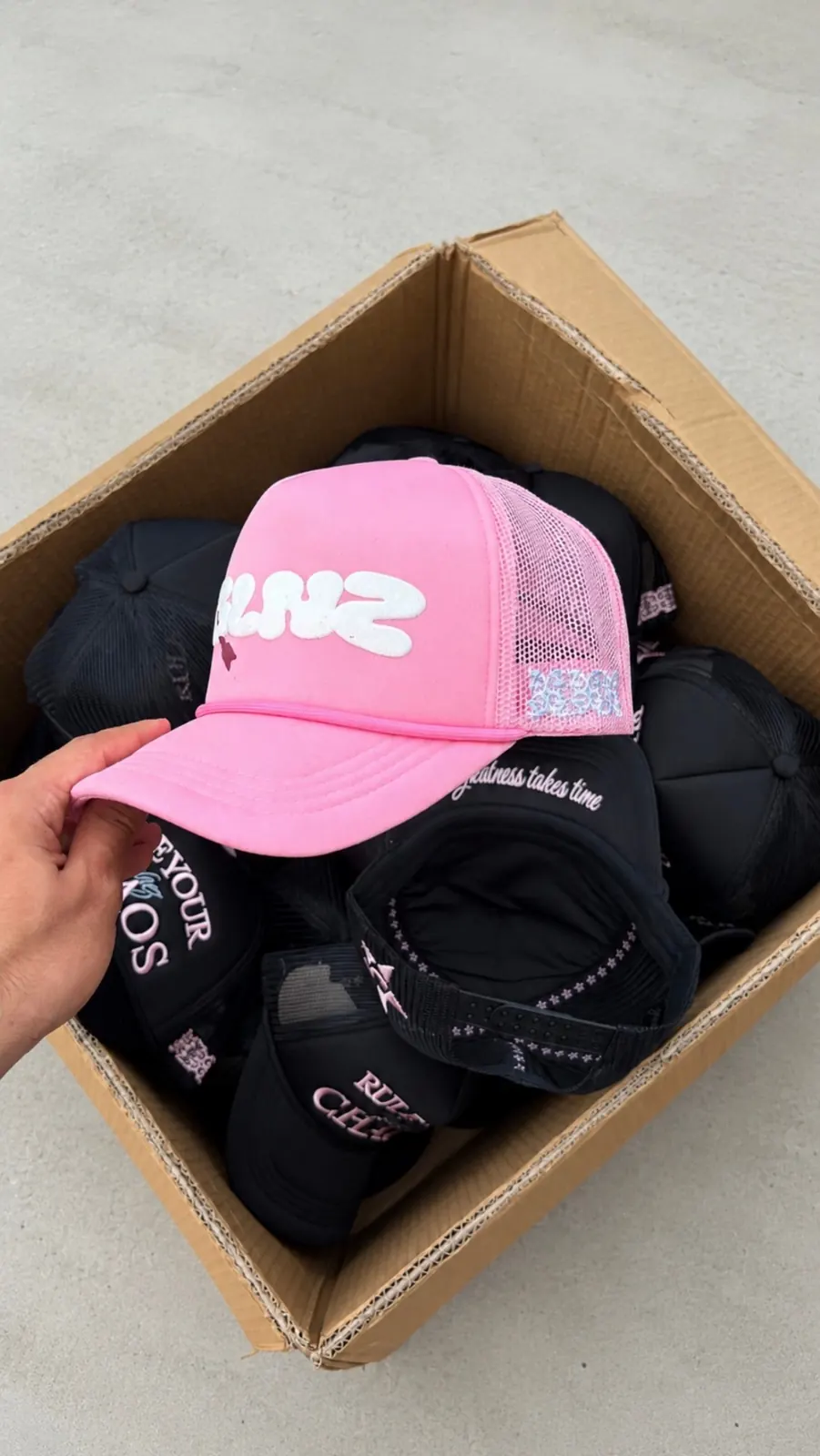 Pink GALNEZ cap held above a box of black caps